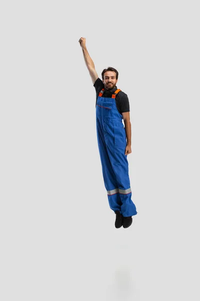 Stripportret van jonge man, mannelijke automonteur in dungarees springen, vliegen omhoog geïsoleerd over witte studio achtergrond. Concept van beroep, grappige meme emoties — Stockfoto