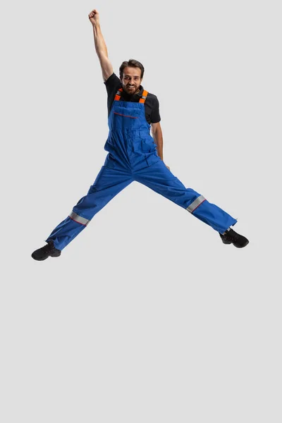 Comic portrait of young man, male auto mechanic in dungarees jumping, flying up isolated over white studio background. Concept of occupation, funny meme emotions — Stock Photo, Image