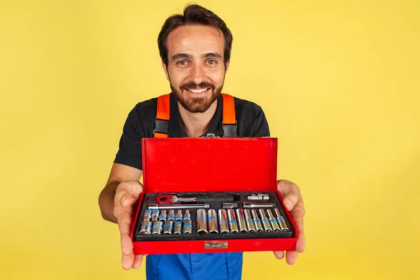 Halblanges Porträt eines lächelnden jungen bärtigen Mannes, eines Automechanikers oder Installateurs in blauer Latzhose, isoliert über gelbem Studiohintergrund. — Stockfoto
