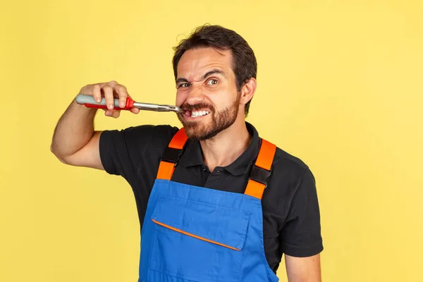 Halblanges Porträt eines lächelnden jungen bärtigen Mannes, eines Automechanikers oder Installateurs in blauer Latzhose, isoliert über gelbem Studiohintergrund. — Stockfoto