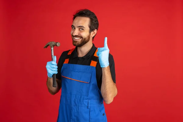 Lustiger junger bärtiger Mann, männlicher Automechaniker oder Installateur in blauer Latzhose vor rotem Studiohintergrund. Konzept der Gesichtsbewegungen — Stockfoto