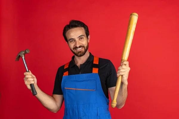 Lächelnder junger bärtiger Mann, männlicher Automechaniker oder Installateur in blauer Latzhose mit Werkzeug vor rotem Studiohintergrund. Konzept der Gesichtsbewegungen — Stockfoto