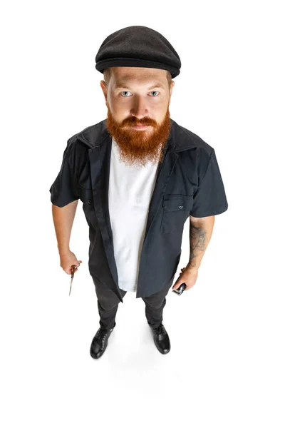 Вид на рыжего бородатого человека, парикмахера или стригущего волосы в черной кепке, выделенный на белом фоне студии. Холдинг его рабочего оборудования. — стоковое фото