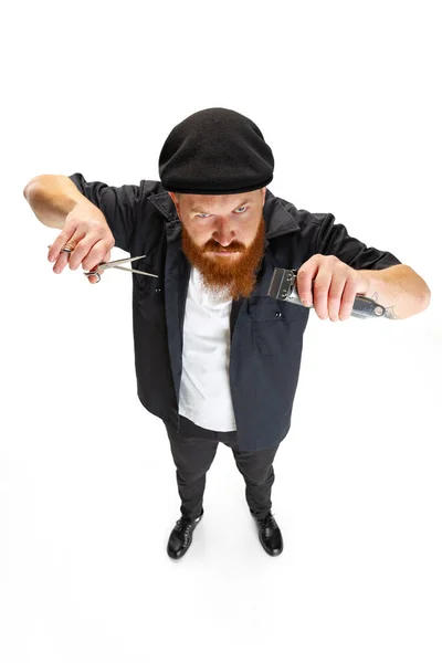 Vista de alto ângulo do homem barbudo vermelho, barbeiro ou cortador de cabelo em boné preto isolado no fundo do estúdio branco. Segurando seu equipamento de trabalho. — Fotografia de Stock