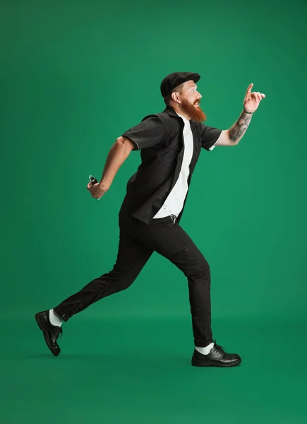 Retrato de hombre de barba roja divertido elegante, peluquero o cortador de pelo corriendo aislado sobre fondo de estudio verde. Sosteniendo su equipo de corte. — Foto de Stock