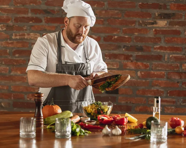 Young Caucasian red-bearded man, chef cooking fresh vegetable salad in cafe, restaurant kitchen. Concept of a correct, healthy diet.