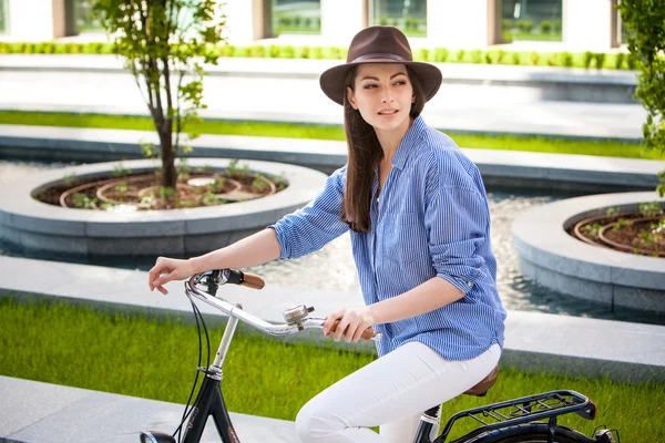 Dość uśmiechający się dziewczyna w kapeluszu i niebieska sukienka, jazda na rowerze na ulicy podnoszenia nogi — Zdjęcie stockowe