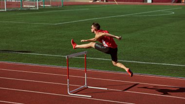 Athlete in red sprinting and jumping over hurdle on green stadium track. Concept of motion, power, motivation for sports campaigns, athletic branding, and dynamic advertising visuals.