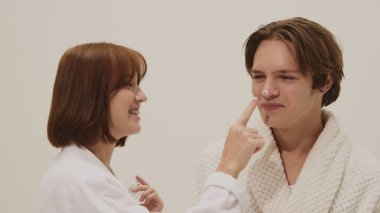 Beyaz bornozlu neşeli kadın gülümseyen erkek burnuna cilt kremi sürüyor. Güzellik ve cilt bakımı endüstrisi, sağlık yaşam tarzı, dermatoloji sağlığı ve reklam kampanyaları kavramı.