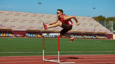 Erkek sporcu, güneşli antrenman sırasında stadyum pistinde engelin üzerinden atlıyor. Kararlılık kavramı, atletik enerji, spor markalaşması, motivasyon ve fitness reklamcılığına odaklanma.