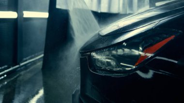 Detaylı stüdyoda arabanın ön tarafı yüksek basınçlı su akışıyla durulanıyor. Temizlik teknolojisi, servis terfisi ve otomotiv bakım reklamı kavramı.