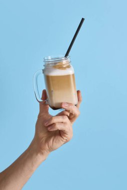 Buzlu latte ve kafe reklamları için mavi arka planda saman ile el ele tutuşan cam bardak. İçecek sunumu kavramı, menü, ambalaj ve pazarlama tasarımı için ürün fotoğrafçılığı.