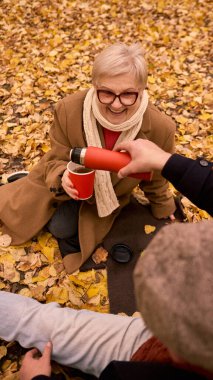 Sonbahar piknik battaniyesinde kırmızı termostan kağıt bardağa sıcak çay dökmek. Yiyecek markalaşması, yaşam tarzı reklamcılığı ve refah içeriği kavramı sıcaklığı ve özeni vurguluyor.