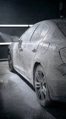 Parlak çamaşır ışıkları altında tamamen kar köpüğüyle kaplı siyah bir araba. Araba bakım ürünleri kavramı, otomobil bakım hizmeti, yol güvenliği farkındalığı, ürün tanıtımı görselleri.