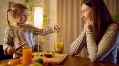 Anne ve kızının neşeli ruh haliyle eğlenceli meyve hazırlıkları sırasında taze portakal suyu masada. İçki görselleri, aile hayatı reklamları, sağlıklı rutinler ve duygusal sıcaklık kavramı..