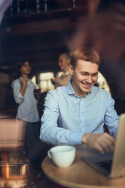 Kafede dizüstü bilgisayarda çalışan ve arka planda sohbet eden bir adam. Esnek çalışma düzenleri, mobil üretkenlik, karışık sosyal iş akışı ve modern kafe ofis kültürü kavramı.