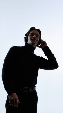 Siyah balıkçı yakalı genç adam kulaklıkla beyaz arka planda müzik dinliyor. Modern yaşam tarzı kavramı, minimal moda tarzı, gençlik kültürü ve günlük teknoloji kullanımı.