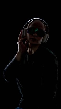 Kafası kazınmış, fütüristik kulaklık takan ve karanlık stüdyoda güneşlik takan genç bir kadın. Kapsamlı teknoloji kavramı, dijital kimlik, kişisel deneyim, sanal arayüzler, modern teknoloji yaşam tarzı.