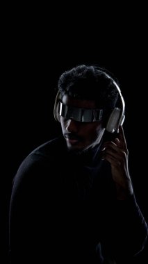 Karanlık stüdyoda fütüristik kulaklık takan ve kulaklık takan bir adam. Kapsamlı medya kontrolü, akıllı ses aygıtları tanıtımı, dijital arayüzler ve modern teknoloji kültürü kavramı.