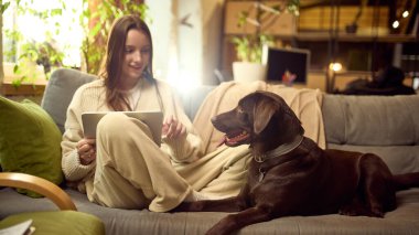 Modern oturma odasında kanepede köpeğiyle tablet kullanan bir kadın. Uzak çalışma rutini kavramı, günlük dijital görevler, çevrimiçi hizmet kullanımı ve evcil hayvan dostu ev ortamı.