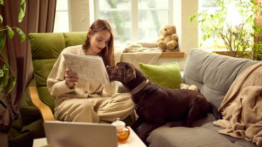 Oturma odasında köpeğiyle kanepede gazete okuyan bir kadın. Günlük rutin kavramı, çevrimdışı medya alışkanlıkları, iş hayatı dengesi, sakin ev atmosferi ve günlük iç istikrar.