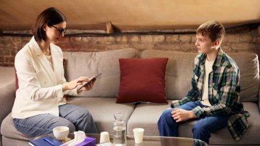 Genç çocuk, kadın terapistle psikoloji seansına tablet kullanarak katılıyor. Profesyonel danışmanlık, ergenlik değerlendirmesi, gençlik destek programları ve modern terapi uygulamaları kavramı.