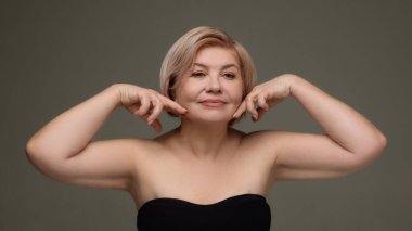 Olgun bir kadın çene hattını işaret ediyor. Yaşlanma karşıtı cilt bakımı. Tedavi reklamını kaldırma, yüz hatları tanıtımı, kozmetik markalaşma, elliden fazla kadın için güzellik kampanyası.