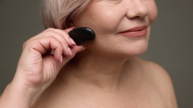 Yanağında gua sha taşı olan olgun bir kadın. Yaşlanma karşıtı yüz masajı, cilt bakım ürünü tanıtımı, cilt bakımı markalaşması, güzellik kampanyası.