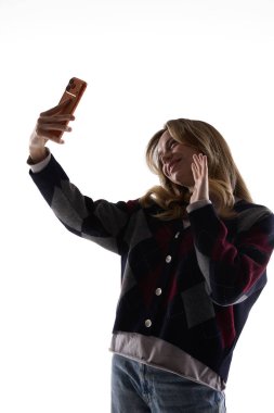 Beyaz arka planda izole edilmiş akıllı telefonlu selfie çeken genç bir kadın. Kişisel markalaşma içeriği oluşturma, sosyal medya tanıtımı ve canlı video yayını kavramı.