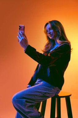 Akıllı telefonlu selfie çeken genç bir kadın portakal renkli arka planda taburede oturuyor. Kişisel markalaşma kavramı, sosyal medya profili güncellemesi, çevrimiçi ağ oluşturma ve dijital iletişim.