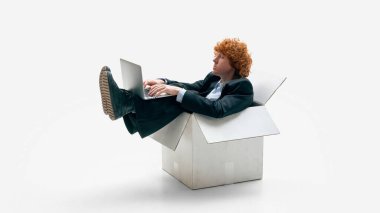 Karton kutunun içinde oturan bir iş adamı beyaz stüdyo arka planı üzerinde çalışıyor. Başlangıçta minimalizm kavramı, sınırlı kaynaklar, önyükleme ve esnek çalışma alanı kültürü.