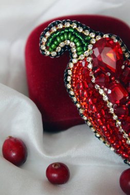 Beyaz ipeğin üzerinde kırmızı biber şeklinde, boncuklarla işlenmiş bir broş ve kırmızı kadife bir kutu bulunur. Kırmızı kızılcıklar beyaz ipeğe saçılmış..