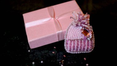 Pembe örgü bir şapka şeklinde boncuklu bir broş koyu yeşil mermer arka planda ipek bir kurdeleyle bağlanmış pembe bir hediye kutusunun yanında..