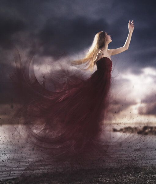 Artistic photography manipulation of a girl flying with a long red dress with a cloudy sky in the sunset. Blonde woman levitate.