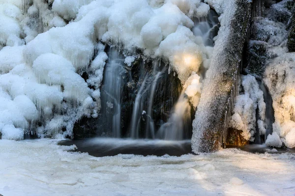 Beautiful frozen waterfall in the winter. Frozen water an ice everywhere. Icy wood log and rocks. Light rays shining through the woods