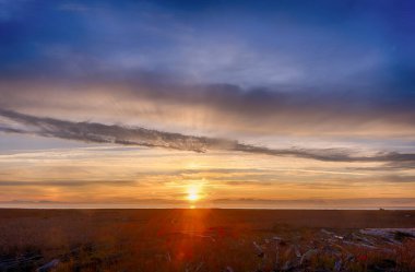 Güzel HDR gün batımı manzarası. Batan güneş bulutlarda güneş ışınları oluşturur. Lens alevi görünüyor. Çim tarlası ve ağaç gövdeleri ön planda. Arka planda dağlar var. 