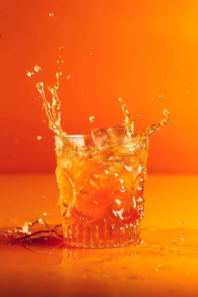 Ice falls into a cut crystal glass with orange liquid and causes a splash on an orange background