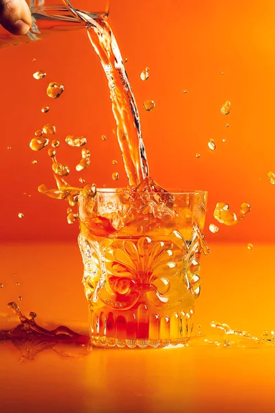 Hand pours liquid into cut crystal glass on orange background and causes a splash.