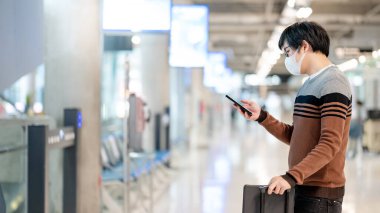 Çantalı Asyalı turist havaalanı terminalinde akıllı telefon kullanarak maske takıyor. Yurtdışına seyahat ederken koronavirüs (COVID-19) salgını önleme. Sağlık farkındalığı ve sosyal uzaklık