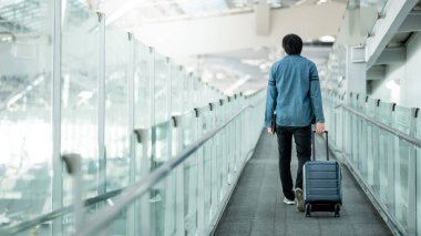 Havaalanı terminalinin girişinde yürüyen yolda bavul ve dijital tablet taşıyan Asyalı turist ya da erkek yolcu. Seyahat sigortası kavramı