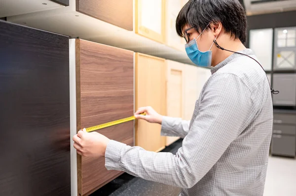 Asian man wearing face mask using tape measure on cabinet panel choosing wooden materials for built-in furniture design. Shopping furniture and house decoration. Home improvement concept