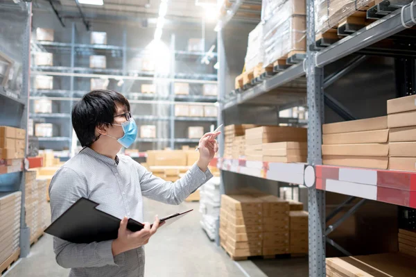 Asian man worker wearing face mask doing stocktaking of product in cardboard box on rack in warehouse by using clipboard. Physical inventory count and preventing the spread of COVID-19 (Coronavirus).