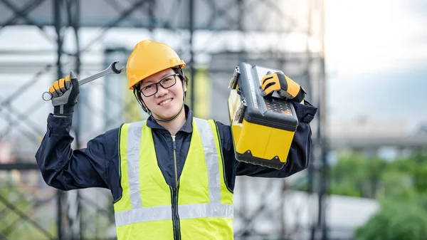 Male Asian mechanic or maintenance worker man wearing protective helmet and reflective vest carrying work tool box and wrench at construction site. Equipment for mechanical engineering project