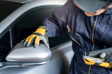 Asyalı oto tamircisi dijital tablet tutuyor. Arabanın kanat aynasını kontrol ediyor. Otomotiv endüstrisinde çalışan makine bakım mühendisi. Otomobil bakım ve onarım kavramı