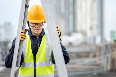 Güvenlik kaskı ve yansıtıcı elbisesi olan Asyalı tamirci inşaat alanında alüminyum merdiveni ayarlıyor. İnşaat mühendisliği, mimarlık, inşaat ve inşaat hizmeti kavramları.