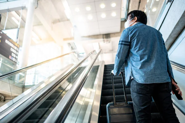 Travel insurance concept. Asian man tourist with face mask carrying suitcase luggage on escalator in airport terminal. Coronavirus (COVID-19) pandemic prevention when travelling.