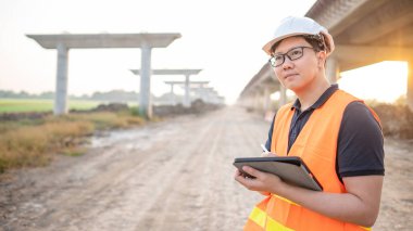 İnşaat alanında proje planlama ve mimari çizimleri için dijital tablet kullanan koruyucu kasklı ve yansıtıcı yelekli akıllı Asyalı işçi ya da erkek inşaat mühendisi..