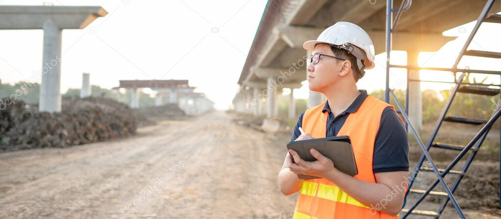 Smart Asian worker man or male civil engineer with protective safety helmet and reflective vest using digital tablet for project planning and checking architectural drawing at construction site.