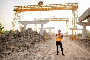 İnşaat alanında proje planlama ve mimari çizimleri için dijital tablet kullanan koruyucu kasklı ve yansıtıcı yelekli akıllı Asyalı işçi ya da erkek inşaat mühendisi..