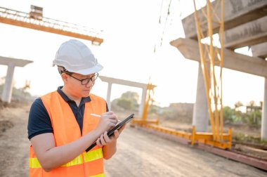 İnşaat alanında proje planlama ve mimari çizimleri için dijital tablet kullanan koruyucu kasklı ve yansıtıcı yelekli akıllı Asyalı işçi ya da erkek inşaat mühendisi..